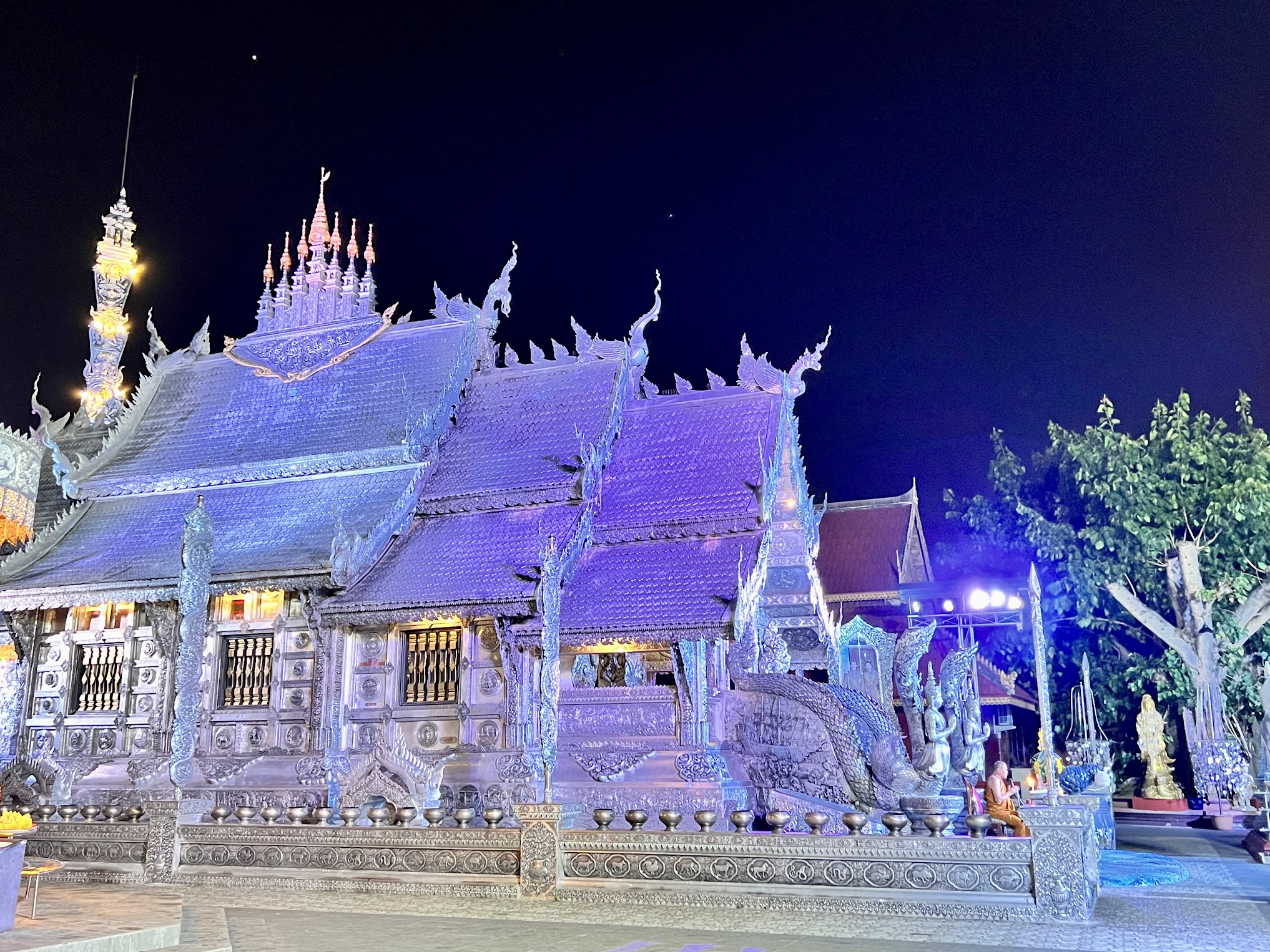 Ornate silver temple viewed from the side and lit up at night with purple lights. There is a monk dressed in orange robes sitting at the front of the temple.