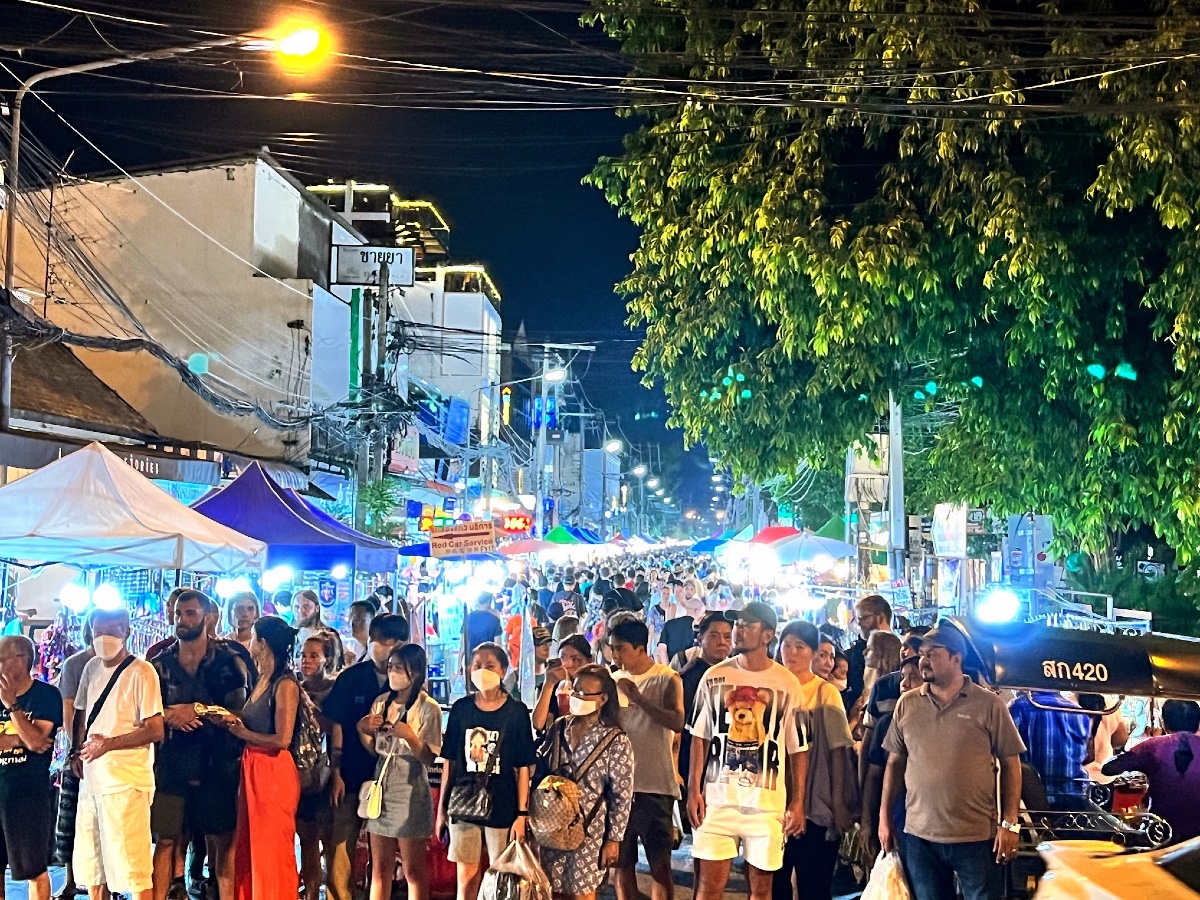 A crowd of people waiting to cross the road from the Chiang Mai nightmarkets. There are stalls lit up with bright lights lining both sides of the road behind the people, and the street - which has been closed to vehicles - is packed with shoppers.