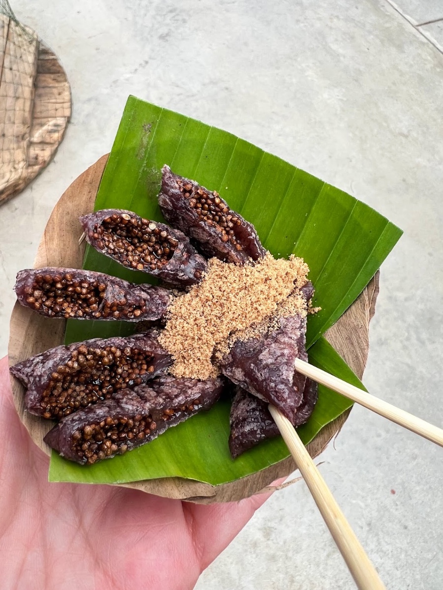 A green banana leaf plate holds seven small pieces of a chia seed pancake sprinkled with cane sugar and speared with bamboo skewers.
