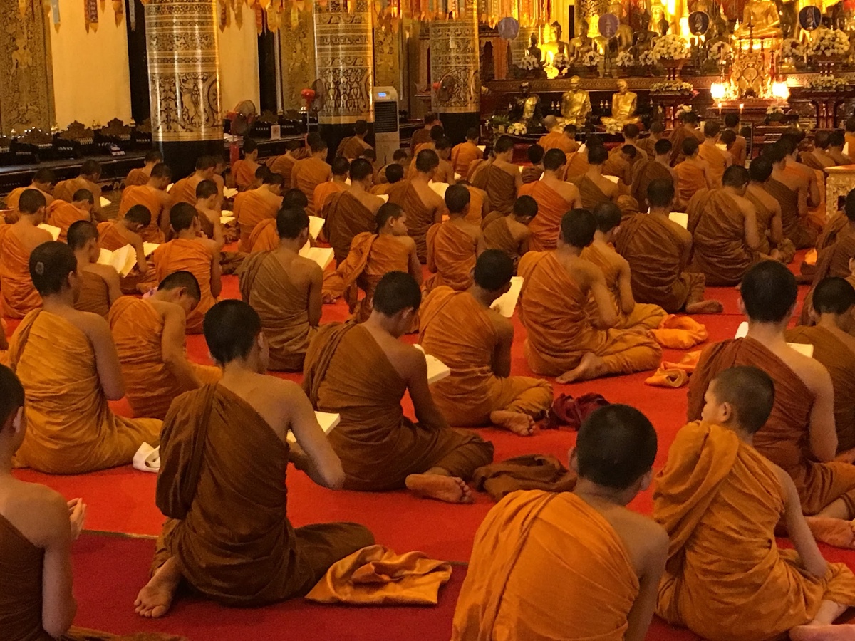 Dozens of young monks robed in orange sit on the floor with their backs to the camera inside a temple.