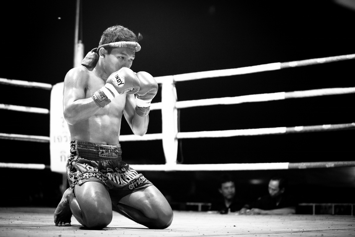 A black and white photo of a Thai Muay Thai fighter kneeling in a boxing ring with his boxing gloved hands raised to protect his face.