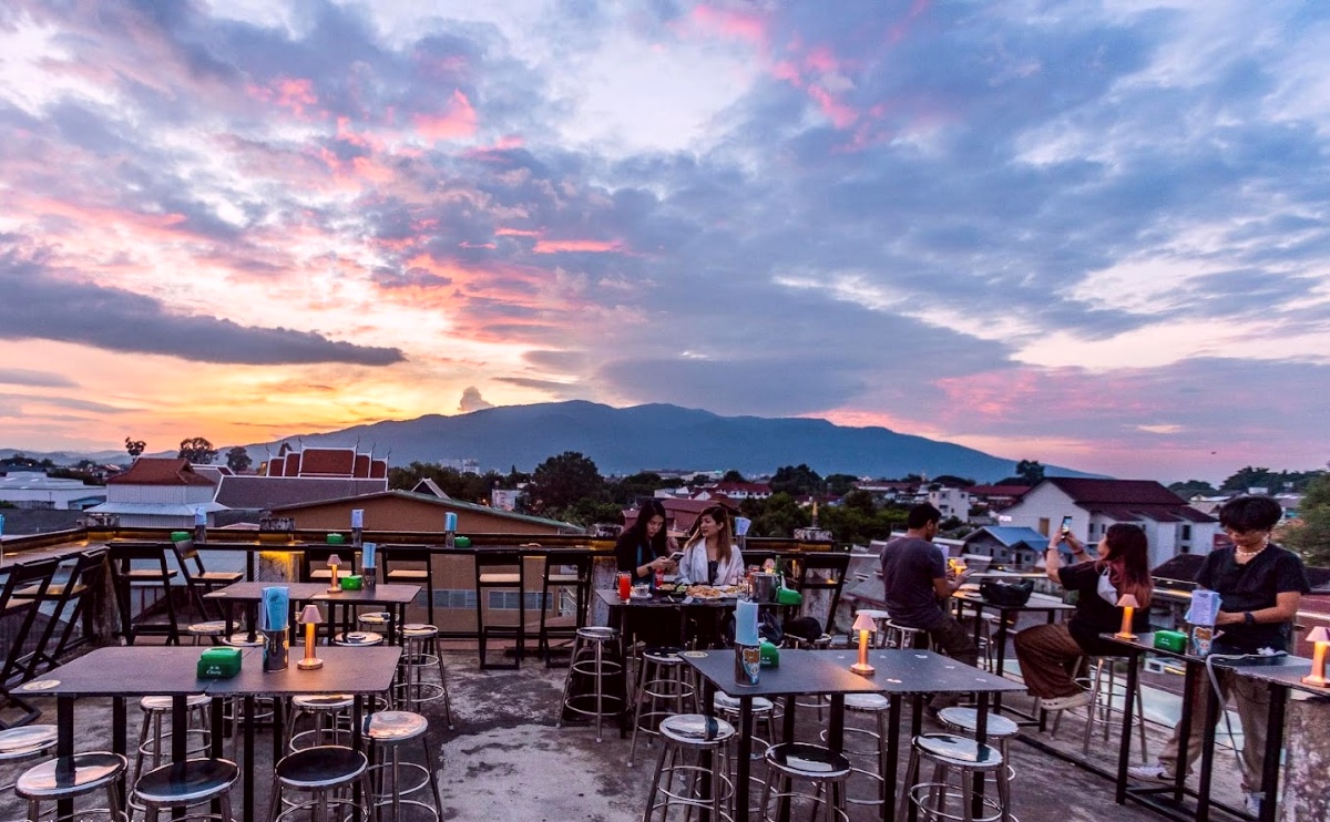 An outdoor rooftop bar scene with several tables and barstools in an open air, uncovered setting. A mountain is visible in the background, and the evening sky is throwing hues of orange, pink, and purple.