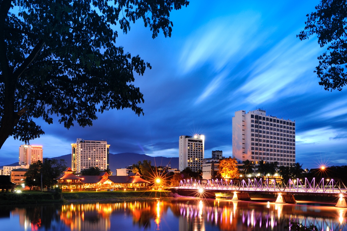 Looking across the Ping River in Chiang Mai at sunset towards the mountains in the background and hotels lit up with orange lights in the middle distance. The lights are reflected on the river's calm surface, and the shadows of trees are in the edges of the foreground. The sky is a deep blue.