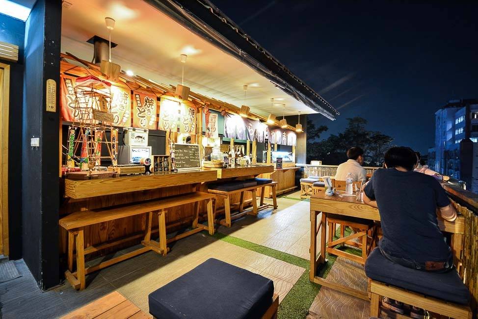 Pale wooden outdoor bar, tables and chairs lit up under a night sky. The scene is casual and calm and the banners hanging behind the bar feature Japanese characters.