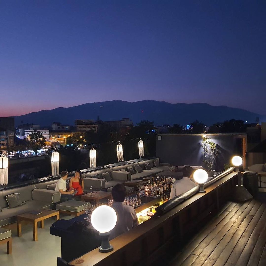 Rooftop bar scene just after sunset with dark purple and blue sky above mountain. Open air bar has a hardwood and cement floors, modular couches and white globe lights.