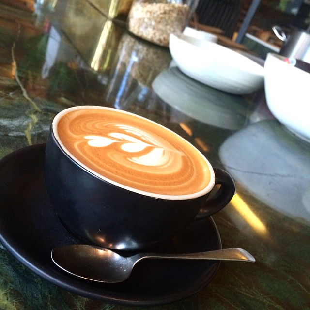 Closeup of a flat white coffee with a swirly decoration on top presented in a black cup and saucer in a Canberra cafe.