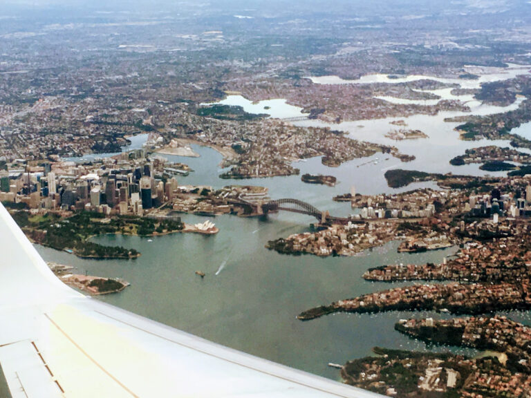 Aerial view from plane window looking white plane wing in foreground and Sydney Harbour in background. Visible are the Sydney Harbour Bridge and the Sydney Opera House.