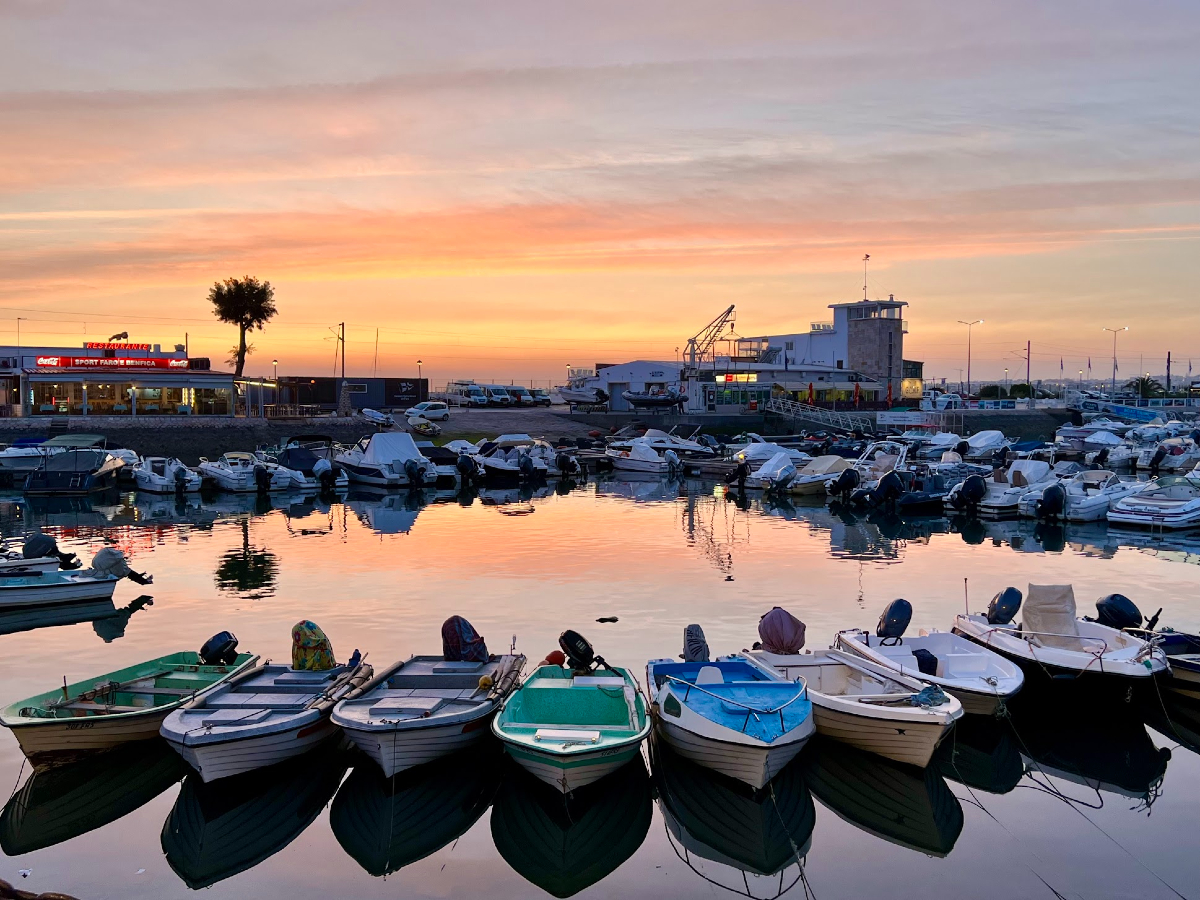 Orange and purplish-blue horizontal streaks across the sky as the sun sets behind the Faro marina. In the background are larger boats, some with masts, and a palm tree, while the foreground has eight small, coloured boats lined up next to one another.