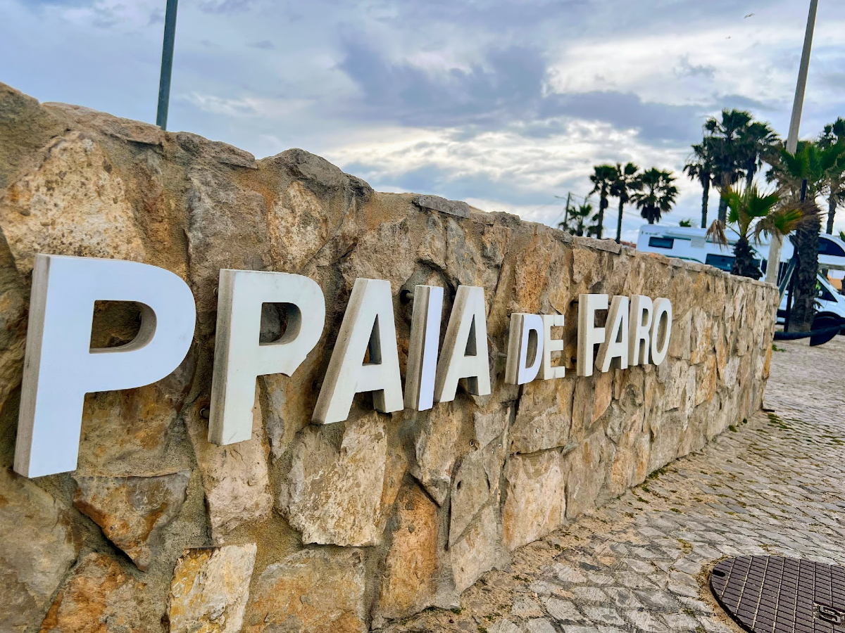 White capitalised lettering reading Praia de Faro on a low stone wall. There are some palm trees in the background.