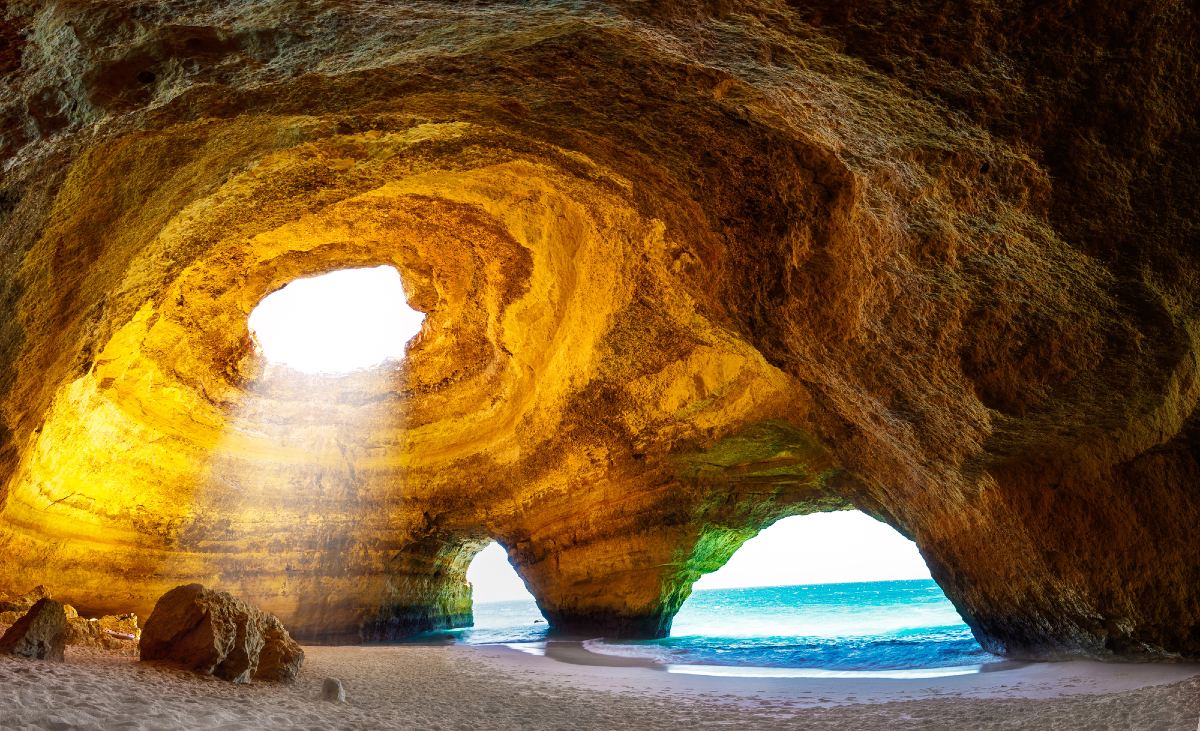 Looking up from inside a sandy-bottomed ocean rock cave with a circular hole in the top through which light is visible, and out two entrances in the side through which the flat, turquoise ocean is visible.