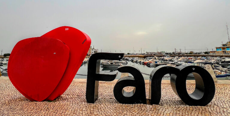 A giant red love heart at the start of giant black lettering reading Faro sitting on the pavement in front of a marina full of small boats.