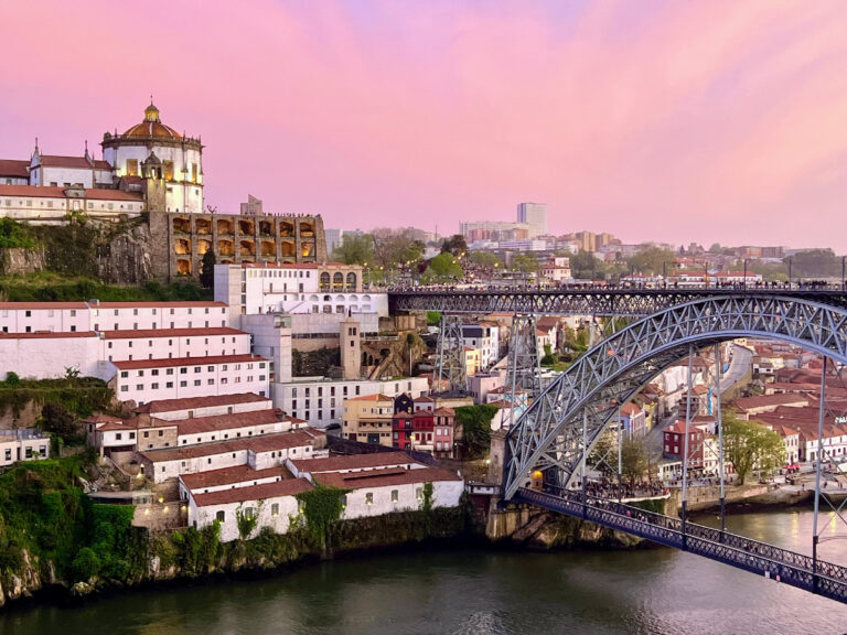 Pink and purple sunset sky behind the white circular building of Serra do Pilar monastery on the hill on the left and the iron arch of Porto's Dom Luis I bridge spanning the Douro River on the right.