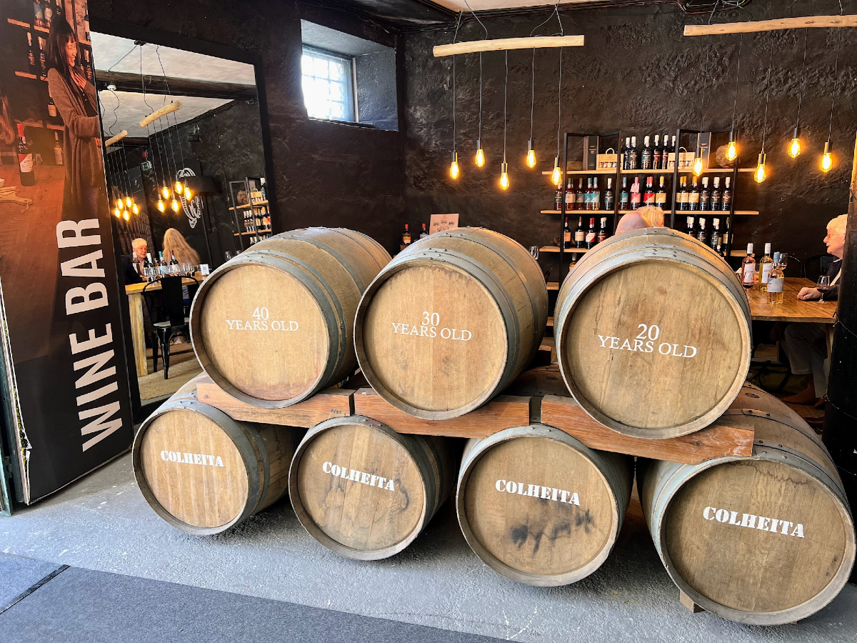 Seven light brown wine barrels lie stacked in the foreground, each marked 20, 30, or 40 years old. In the background are tables and chairs in a wine bar with low hanging lights.