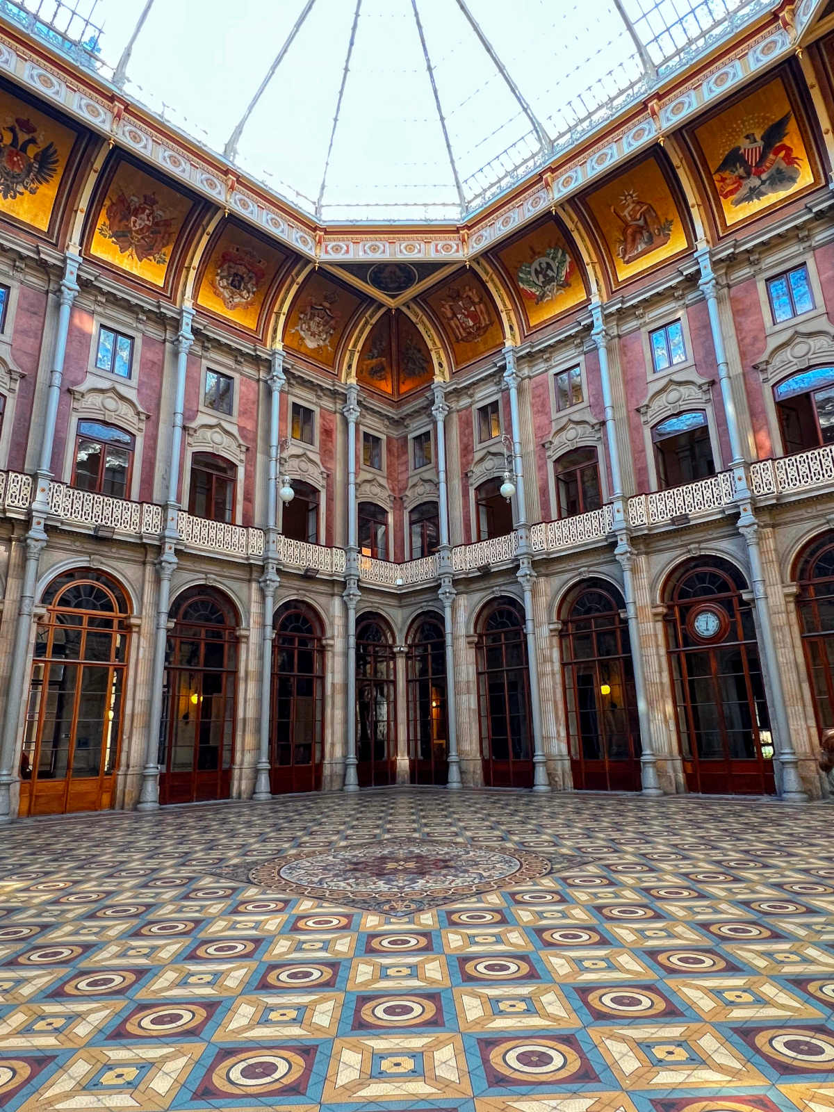 Large central room with domed glass ceiling, ornately tiled floor, and gold pictures around the tops of the walls.