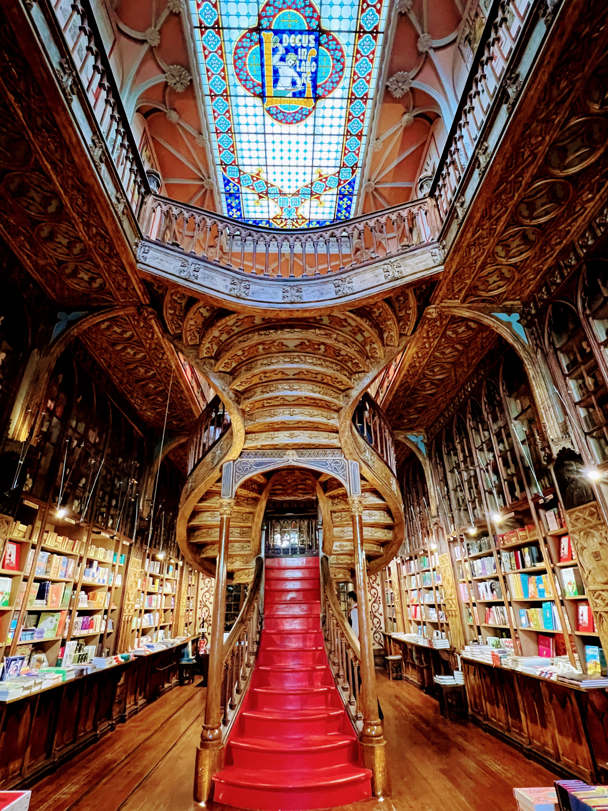 A wide angle view inside the Livraria Lello bookshop. In the centre is a flight of shiny red stairs leading up to the second floor, flanked by intricately carved wooden banisters, the underside of the stairs as they wrap around, and the surround of the stained glass ceiling. The walls are covered in bookshelves.