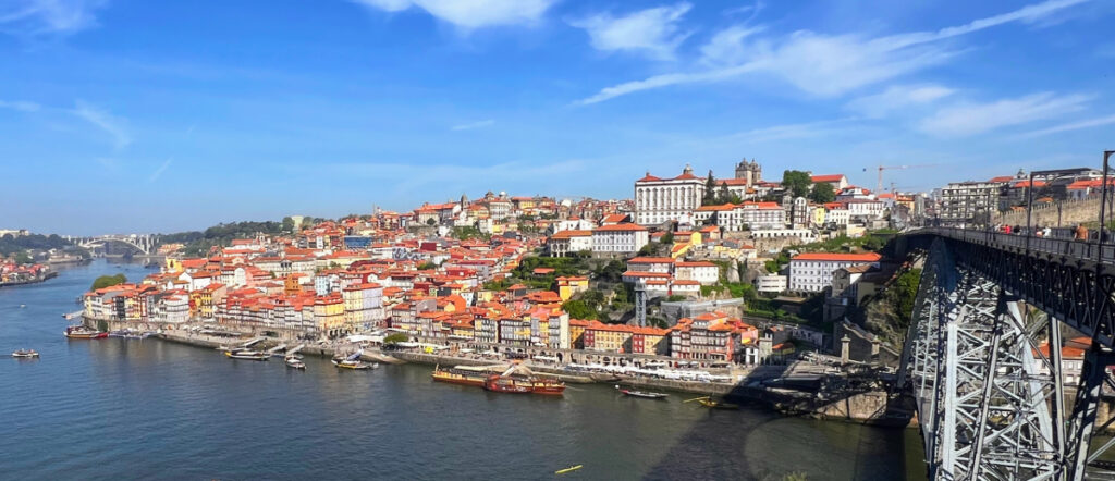 Wide angle view from the top of a bridge looking down across the Douro River to the orange roofs of the city of Porto, Portugal. There are several small boats on the river and the sky above is mid blue with a few wispy white clouds.