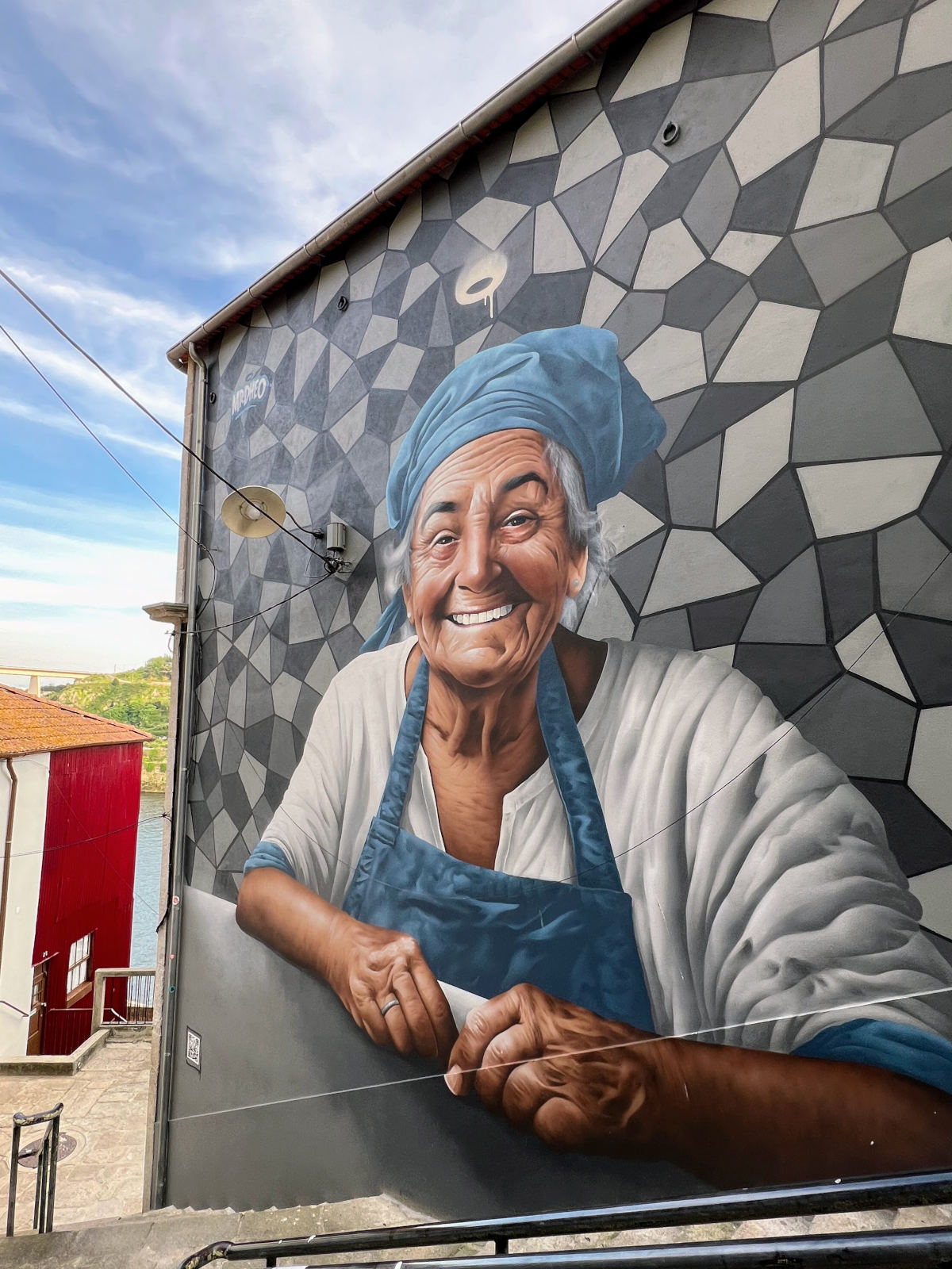 Full wall mural featuring the head and shoulders of an elderly woman smiling broadly. She is wearing a white long sleeved shirt with the sleeves pushed up, a blue apron and blue headscarf. The background is black, white, and grey geometric shapes.