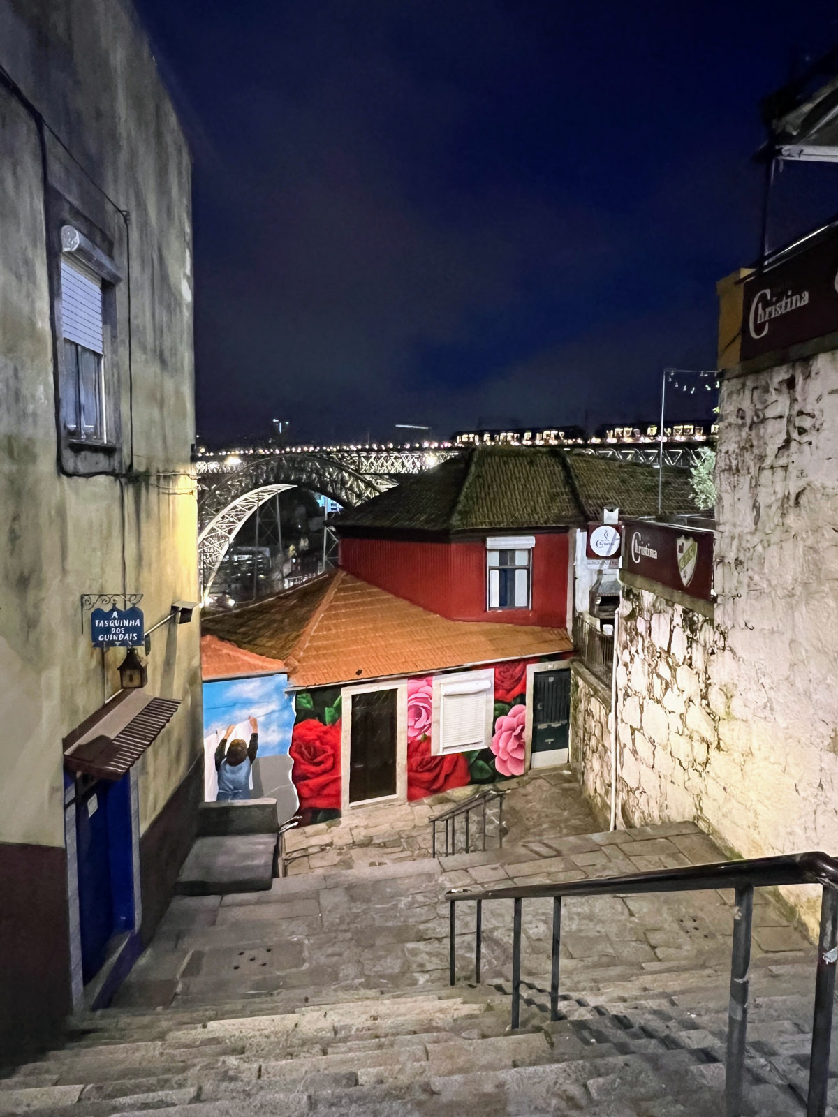 Night view looking down several sets of outdoor stone steps towards mural'd buildings and the lit up Dom Luis I Bridge in Porto in the background.