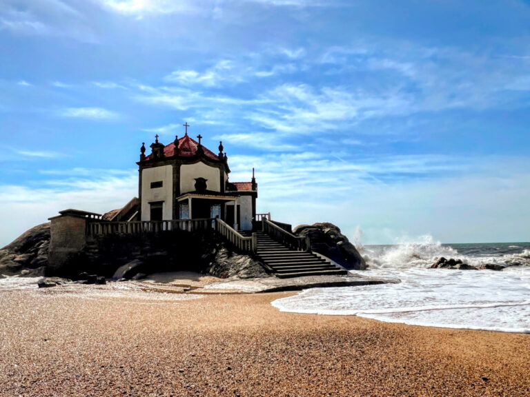 A small, white hexagonal chapel sits on a rocky outcrop at the edge of the ocean. Golden sand in the foreground, and a bright blue sky.