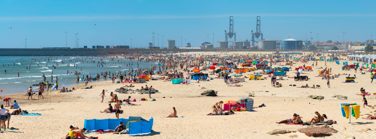 A landscape scene of a wide sandy beach filled with sunbathers. There are cranes in the background.