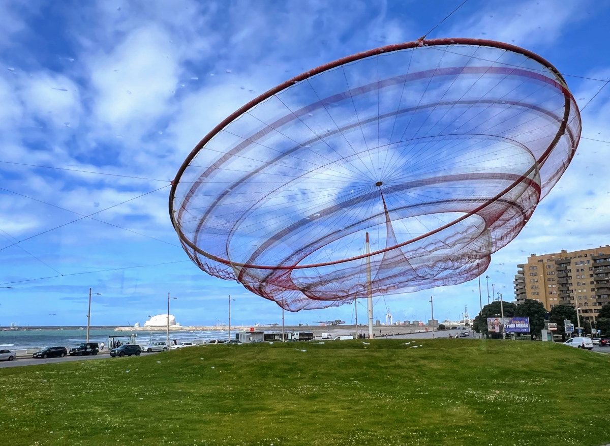 A giant red seafood catching net suspended by cables and flowing in the breeze above a grassy field.