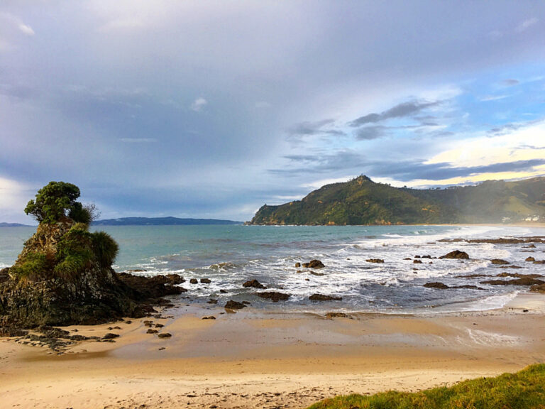 Landscape photo of a beach with golden sand in the foreground, a rock with shrubbery on the ocean's edge, and the sea and rocky headland in the background.