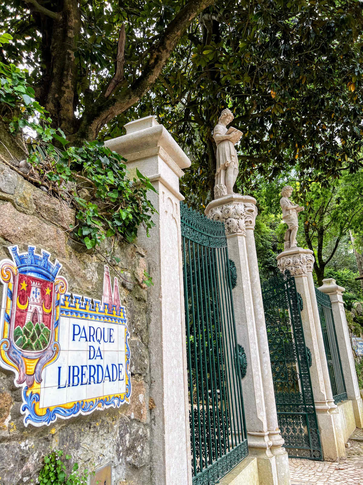 Portrait image of a tall stone wall with green wrought iron gates and an ornate and colourful sign reading Parque da Liberdade