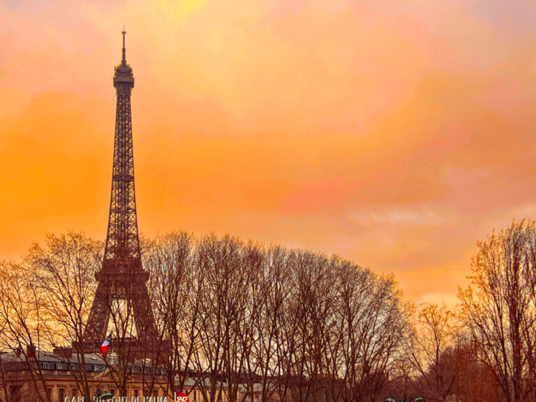 Landscape image of the Eiffel Tower against an orange sky with trees in the background. A very small French flag flies in the foreground.