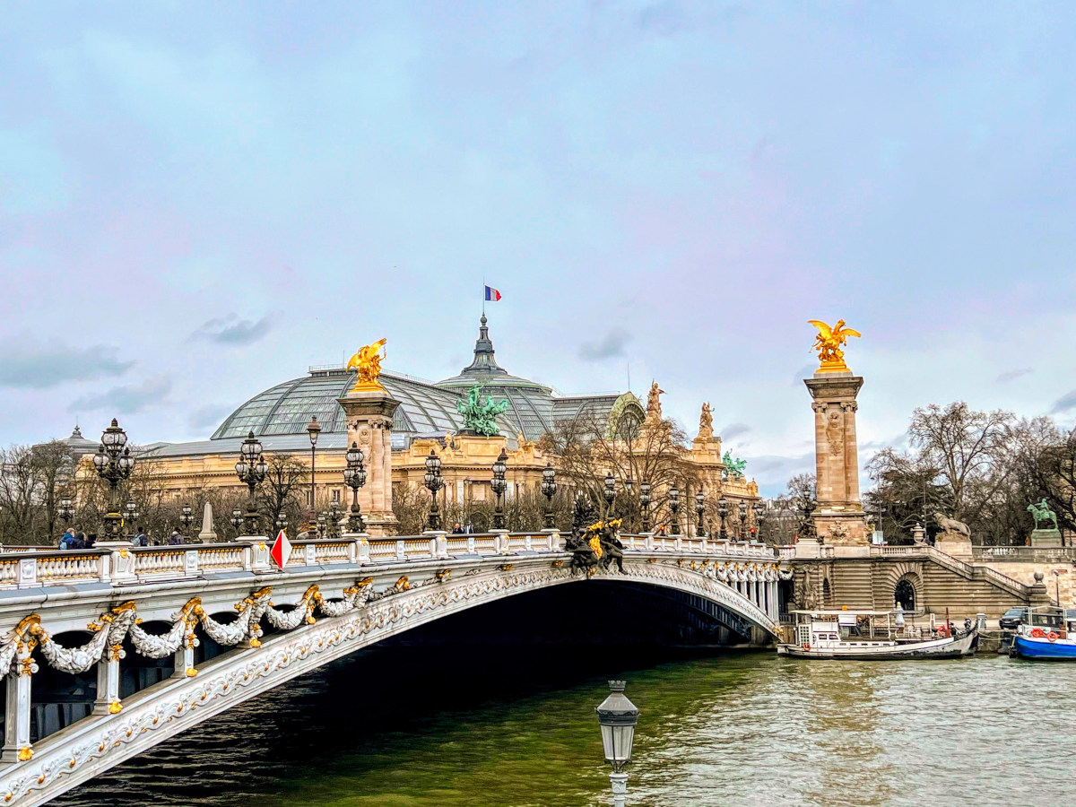 Landscape view of the ornate, single arch Alexandre Bridge III in Paris, complete with gold statues atop stone plinths at each corner. The bridge spans the Seine river with a couple of boats on it.