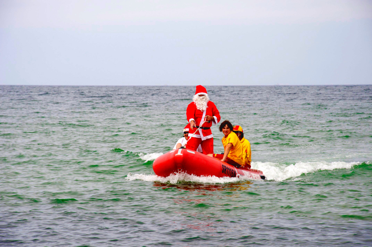 Santa Claus standing on a red inflatable surf lifesaving dinghy as it arrives over the ocean.