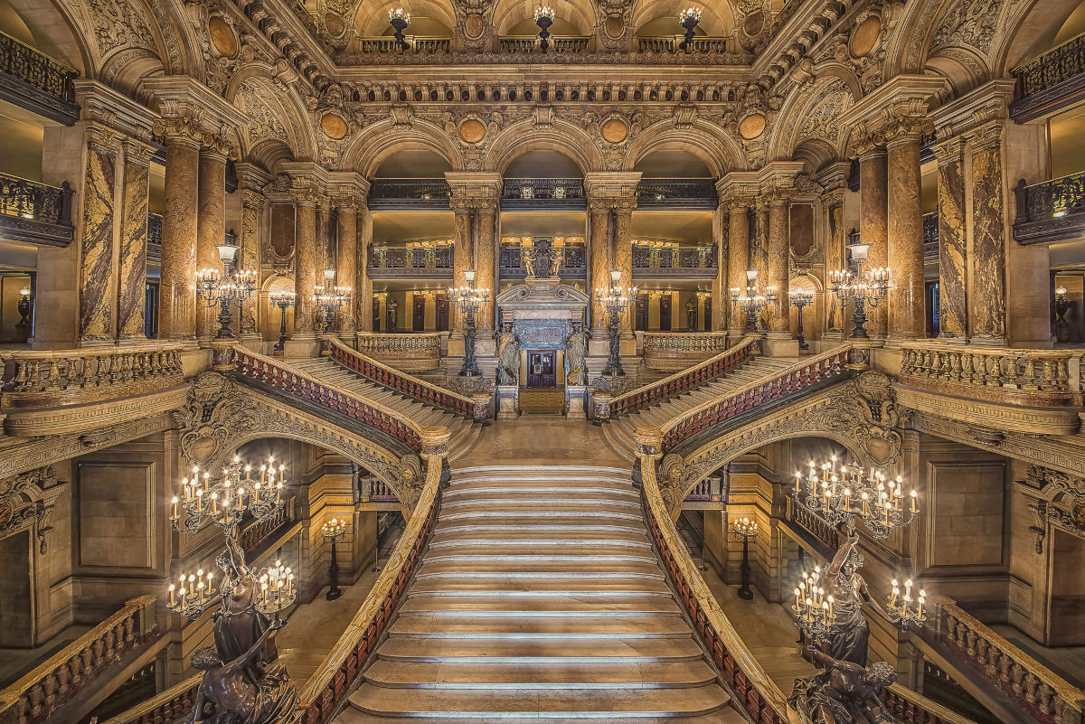 Landscape view of a wide, forked, opulent staircase inside an equally grand and gold hall.