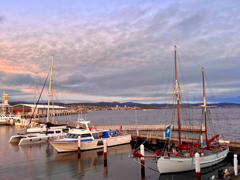 Landscape harbour scene with several small masted boats in the foreground and pinky orange and blue sunrise colours in the sky.