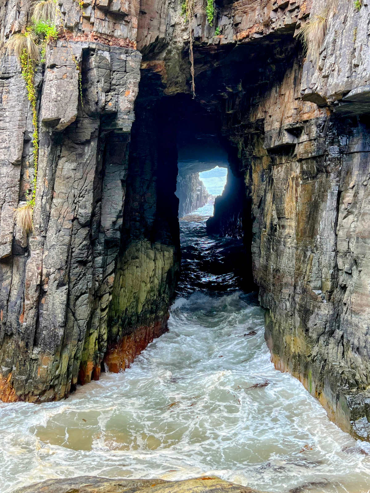 Portrait through a rocky chasm carved by repeatedly crashing sea waves looking out to sea on the far side. White water in foreground.