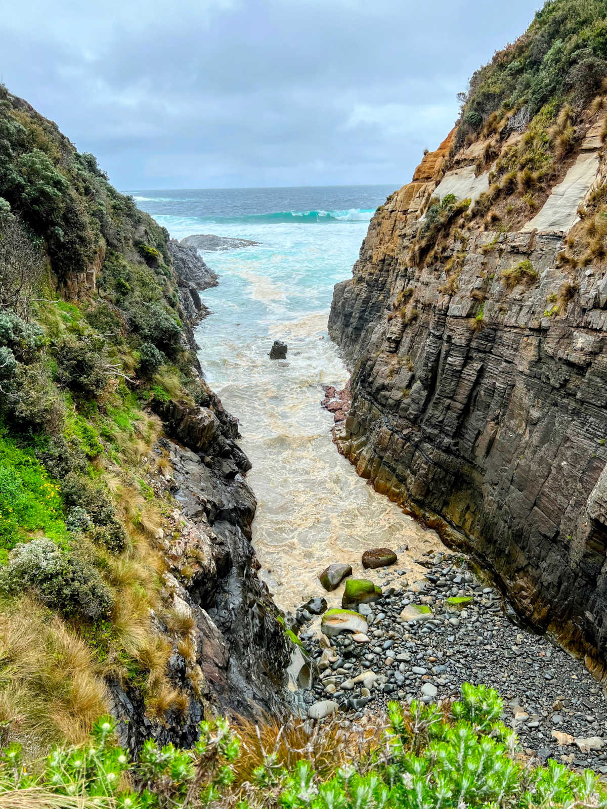 Portrait view from cliff top overhang between rocky sided chasm looking down at sea water and rocks below.