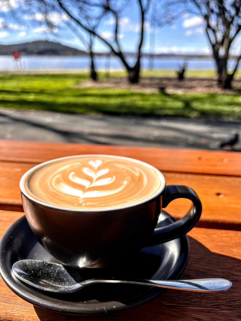 Portrait photo of a coffee in the foreground with tree silhouettes and lake in the background under blue summer sky.