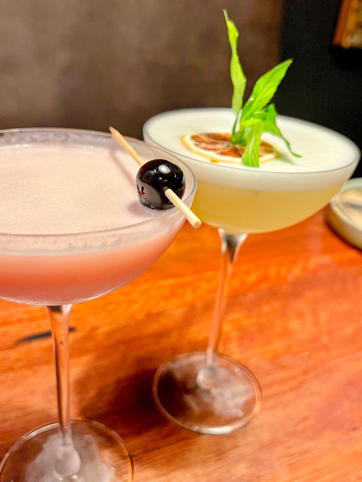 Two martini-style cocktails, one pale pink with a black olive on top, and one pale yellow with greenery on top.